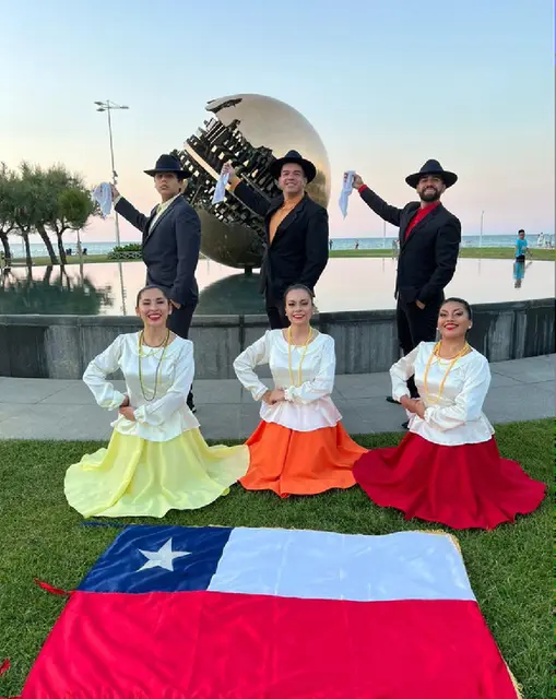 Tres bailarinas con vestidos amarillo, naranjo y rojo y tres bailarines de negro junto a la bandera chilena en Pésaro, Italia